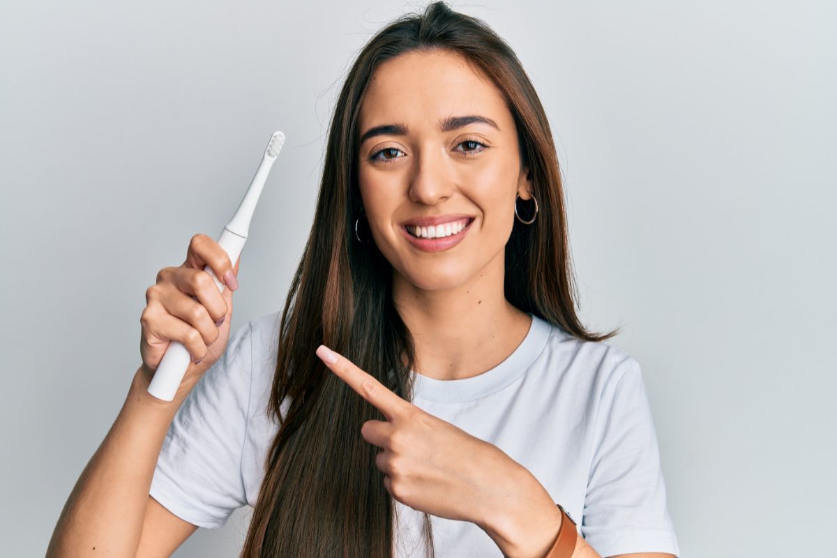 5 New Ways to Improve Your Oral Hygiene Woman holding electric toothbrush pointing at it against white background for dental care website.