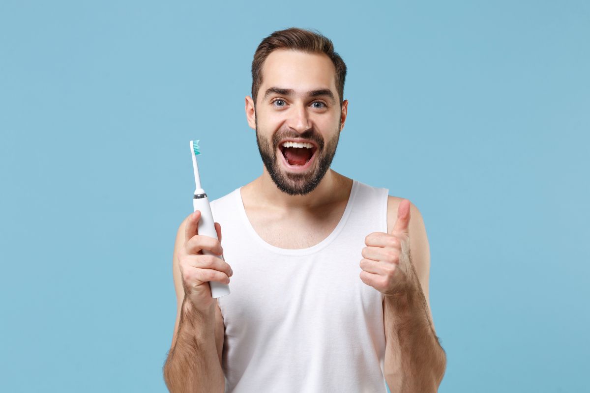 Happy man holding electric toothbrush with thumbs up against blue background