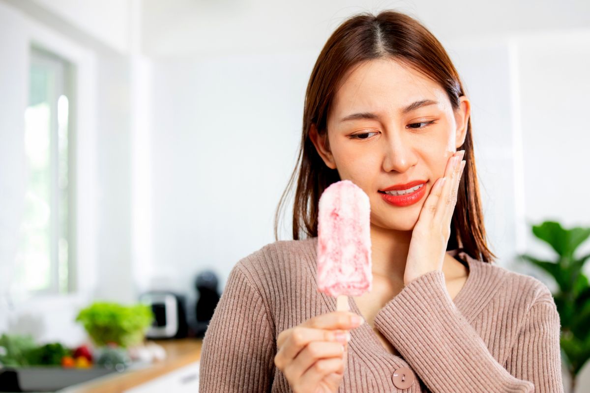 Woman holding ice pack to cheek in bright kitchen, appearing to have tooth pain or swelling.
